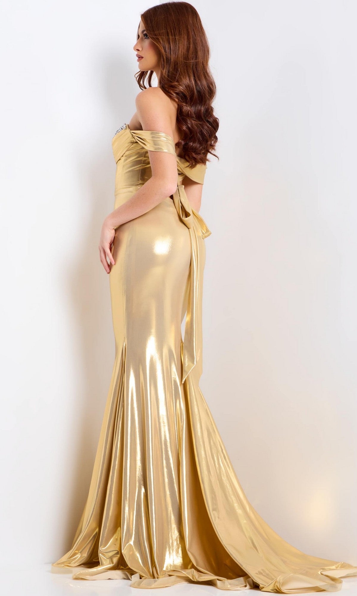 A woman with long brown hair stands in profile wearing the Jovani 47062, a long off-the-shoulder metallic prom dress with a fitted bodice, mermaid silhouette, and slight train.